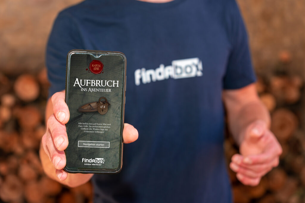 Person in findAbox Shirt zeigt Smartphone digitale Schatzsuche